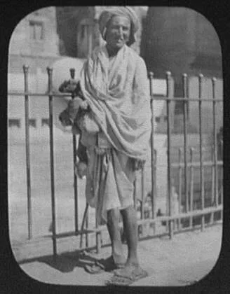 An Indian beggar - standing in front of iron fence, 1895